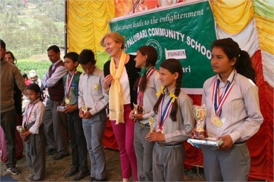 [ai] Students standing in a row at a school event, each holding trophies and medals. An adult is speaking to the group, and a colorful background with a banner that says 'Education leads to enlightenment' is visible.